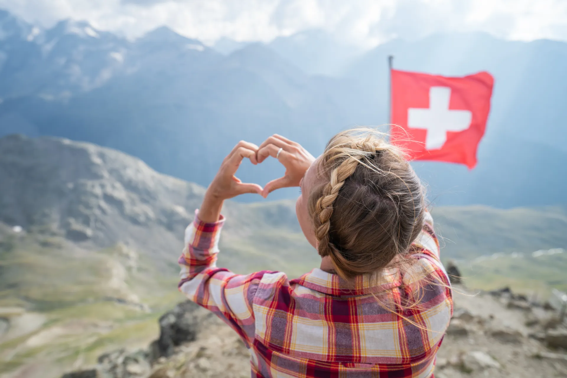 Une femme forme un cœur avec ses mains devant un drapeau suisse et un paysage de montagne.
