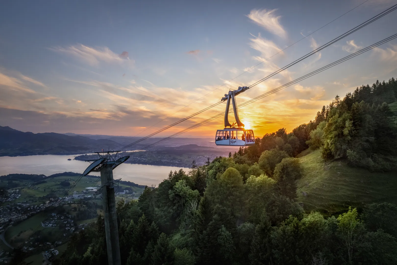 Le téléphérique du Rigi survole des collines boisées au coucher du soleil ; en contrebas s’étend le lac des Quatre-Cantons dans un vaste paysage préalpin.